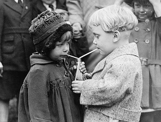 A boy and a girl sharing a drink, each with their own straw in the bottle in 1922.
