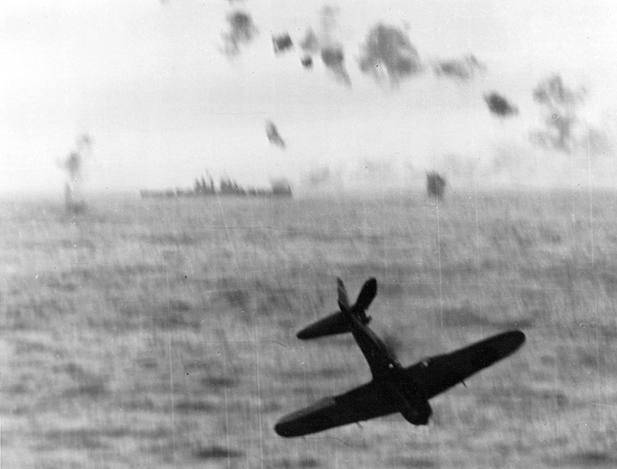 A Japanese Zero Kamikaze crashes into the sea after trying to hit the aircraft carrier USS Essex (CV-9) off Okinawa, May 14, 1945. The vessel in the background is a U.S. Navy Cleveland-class light cruiser.