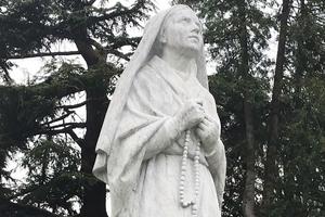 From above: A statue of Bernadette stands in front of the St. Bernadette Museum at the Lourdes sanctuary; the pose of the image reflects her enraptured gaze during each Marian apparition. Pilgrims pray at the Lourdes grotto. The small home where Bernadette lived with her family is now a national monument. The sticks in the fireplace are exactly like those Bernadette was collecting in the woods near the grotto on the day she first met Mary. The crucifix above the fireplace is original. St. Bernadette’s original shawl, wooden shoe and rosary are also on display; the rosary is the one she held during each apparition as she prayed the Rosary with Our Lady.  