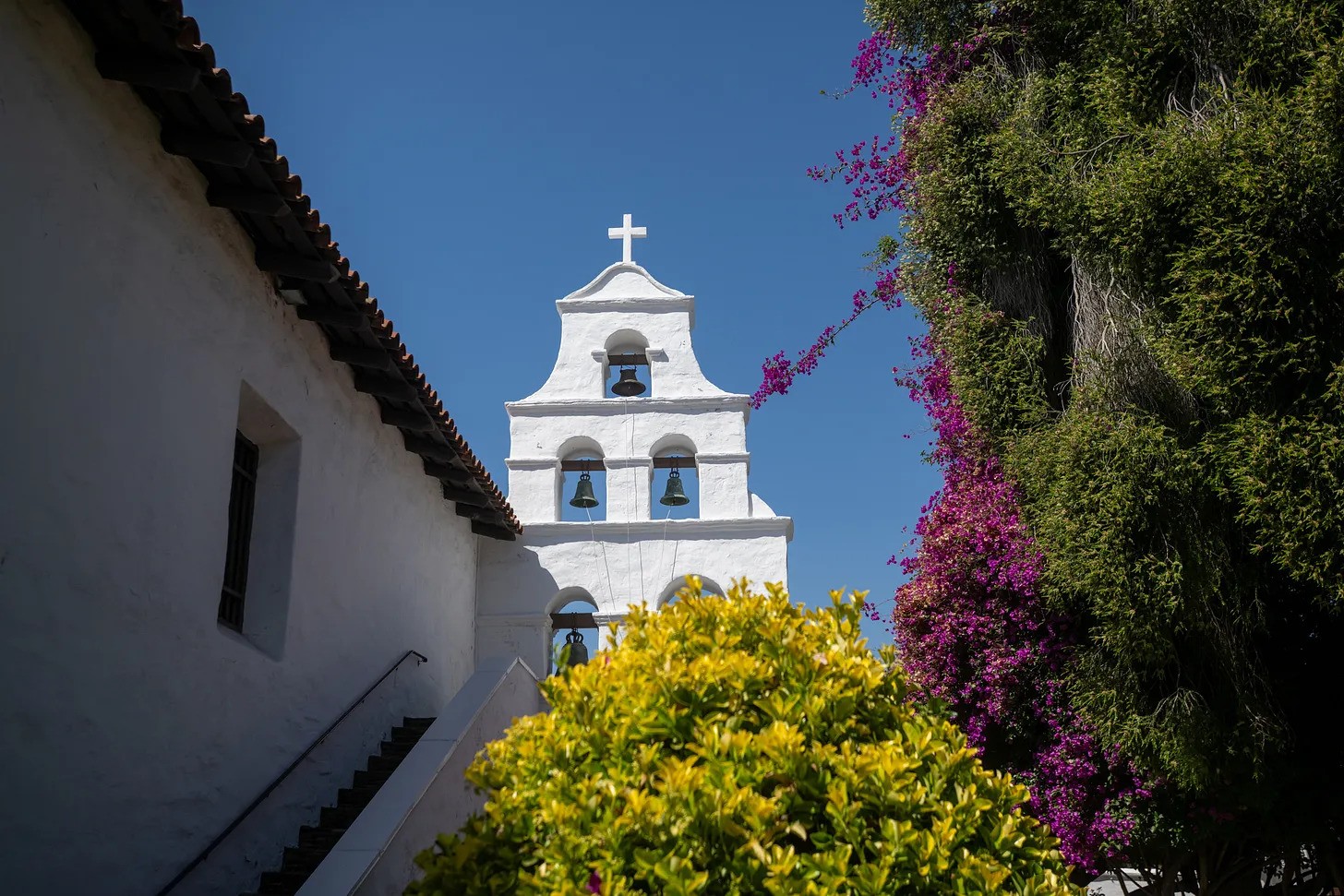The iconic bell tower of Mission San Diego de Alcalá rises beside blooming bougainvillea under a clear blue sky—where faith, history, and beauty meet in California’s first mission.