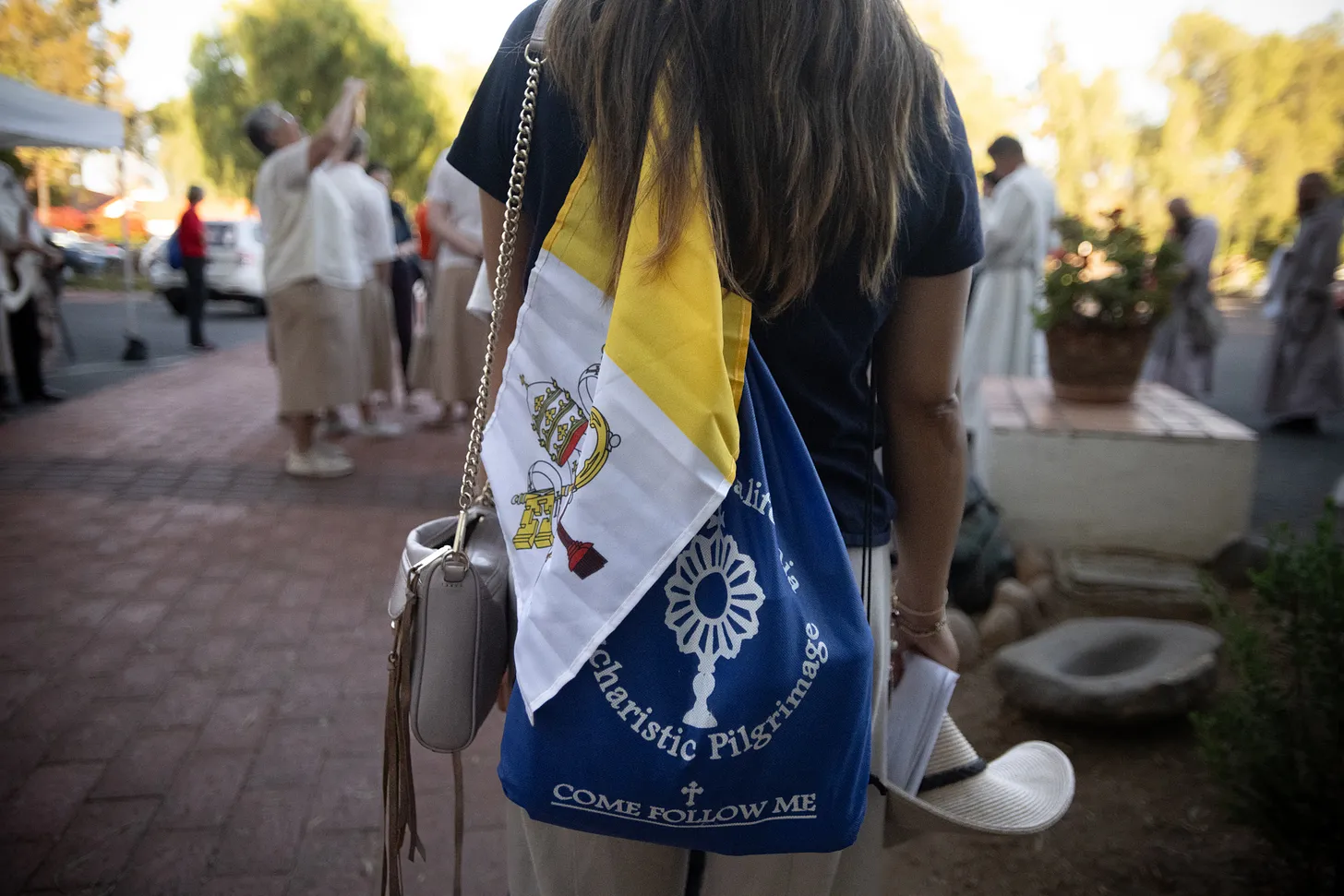A pilgrim wears the official Camino de California Eucharistic Pilgrimage pack, with a Vatican City flag draped from the side, outside Mission San Diego de Alcalá.