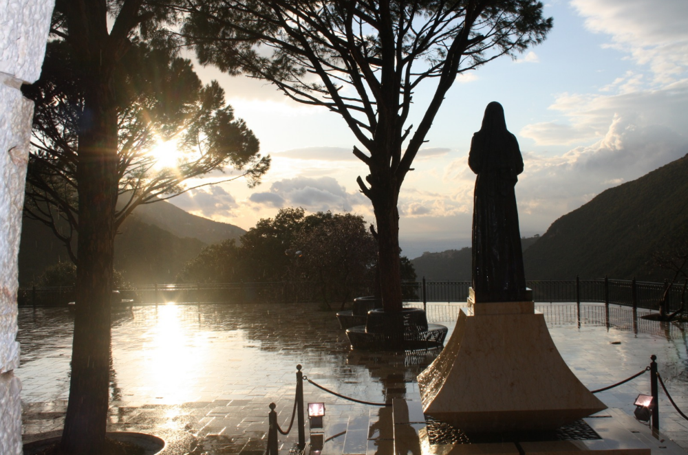 A statue of St. Charbel overlooks a pond and surrounding lush scenery.