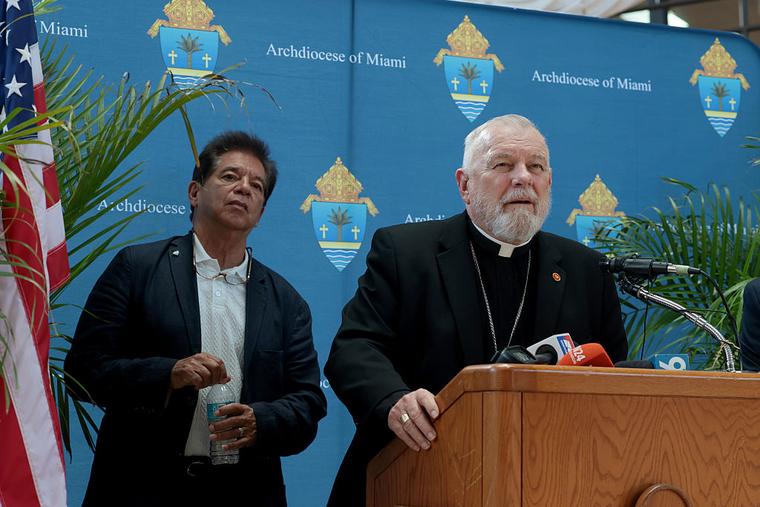 Peter Routsis-Arroyo, Catholic Charities CEO (left), listens as Archbishop Thomas Wenski of Miami speaks during a press conference held at the Archdiocese of Miami Pastoral Center about the cancellation of an $11 million federal contract with Catholic Charities on April 16, 2026, in Miami. For over 60 years, Catholic Charities of the Archdiocese of Miami worked with the federal government to support vulnerable children and families, starting with Operation Pedro Pan, which resettled about 14,000 Cuban children in South Florida.
