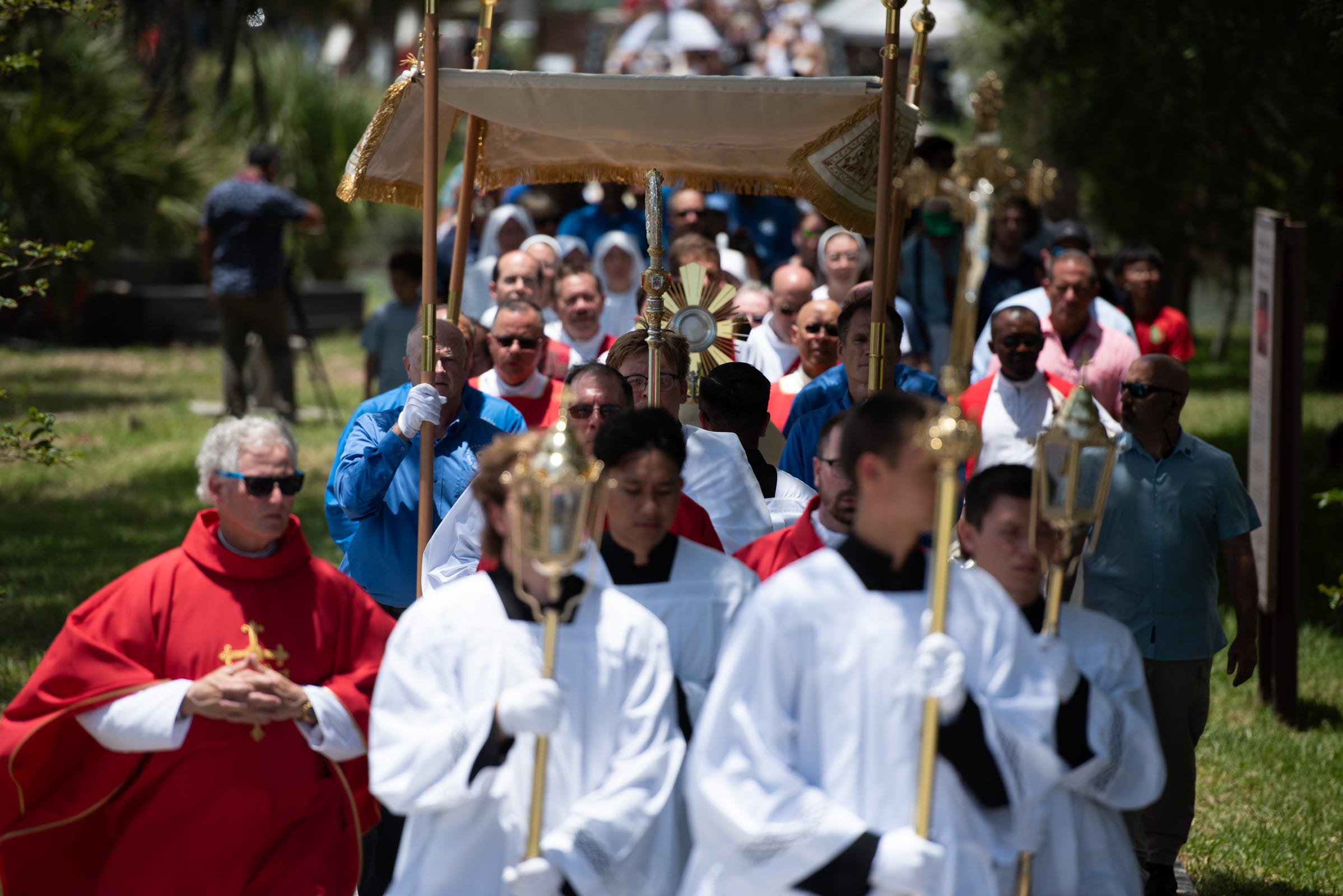 Beneath a canopy and under the midday sun, the Blessed Sacrament is borne through the shrine grounds as clergy, religious, and lay faithful follow in prayer.