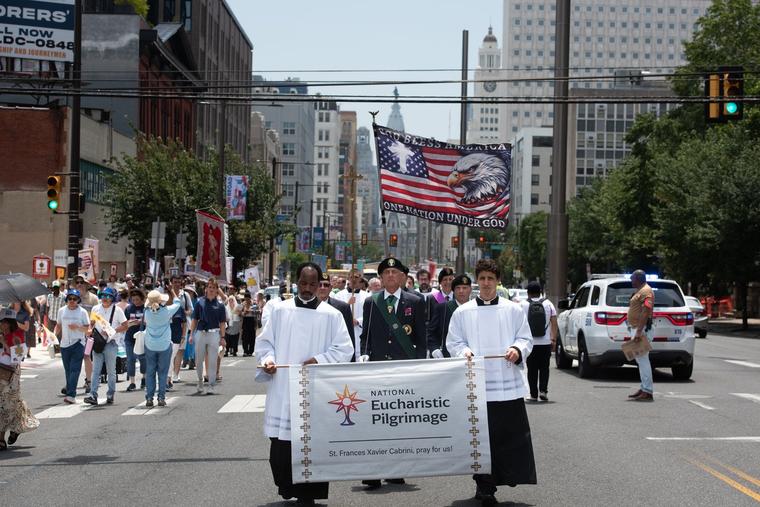 Participants carry the National Eucharistic Pilgrimage banner beneath a flag reading “God Bless America” and “One Nation Under God” during the procession through Philadelphia, Sunday, July 5, 2026. 